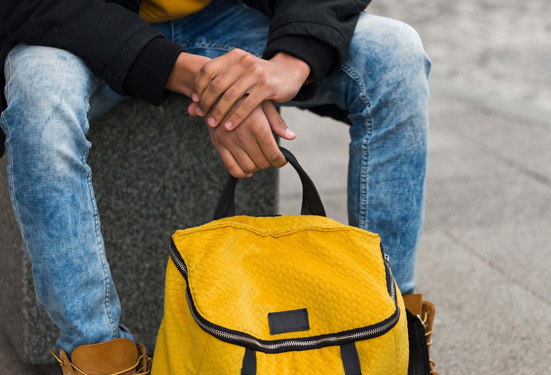 Primo piano di un ragazzo seduto su una panchina che tieni tra le mani uno zaino giallo.
