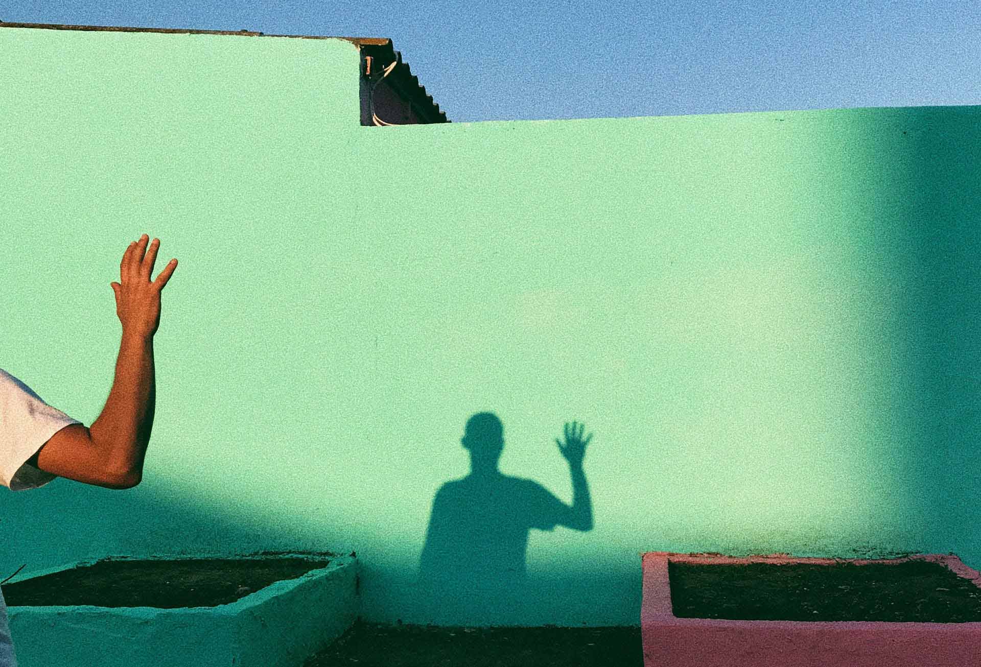 Dettaglio dell'ombra di un ragazzo che saluta, proiettata su un muro verde.